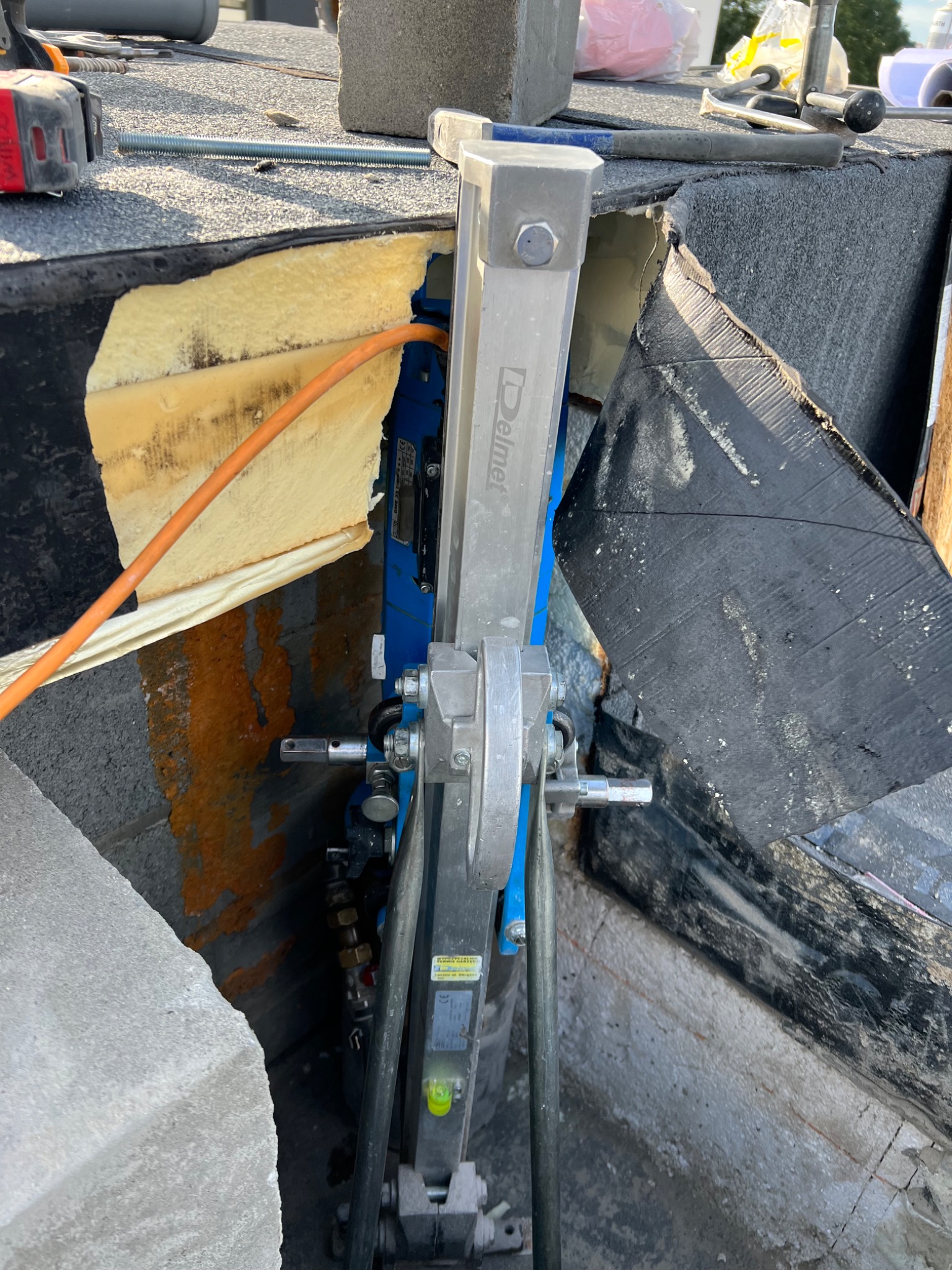 Widok z dołu na rdzeniową wiertnicę Delmet, używaną do wiercenia otworów w dachu pokrytym papą i ocieplonym styropianem. Widoczne przewody i elementy konstrukcyjne.