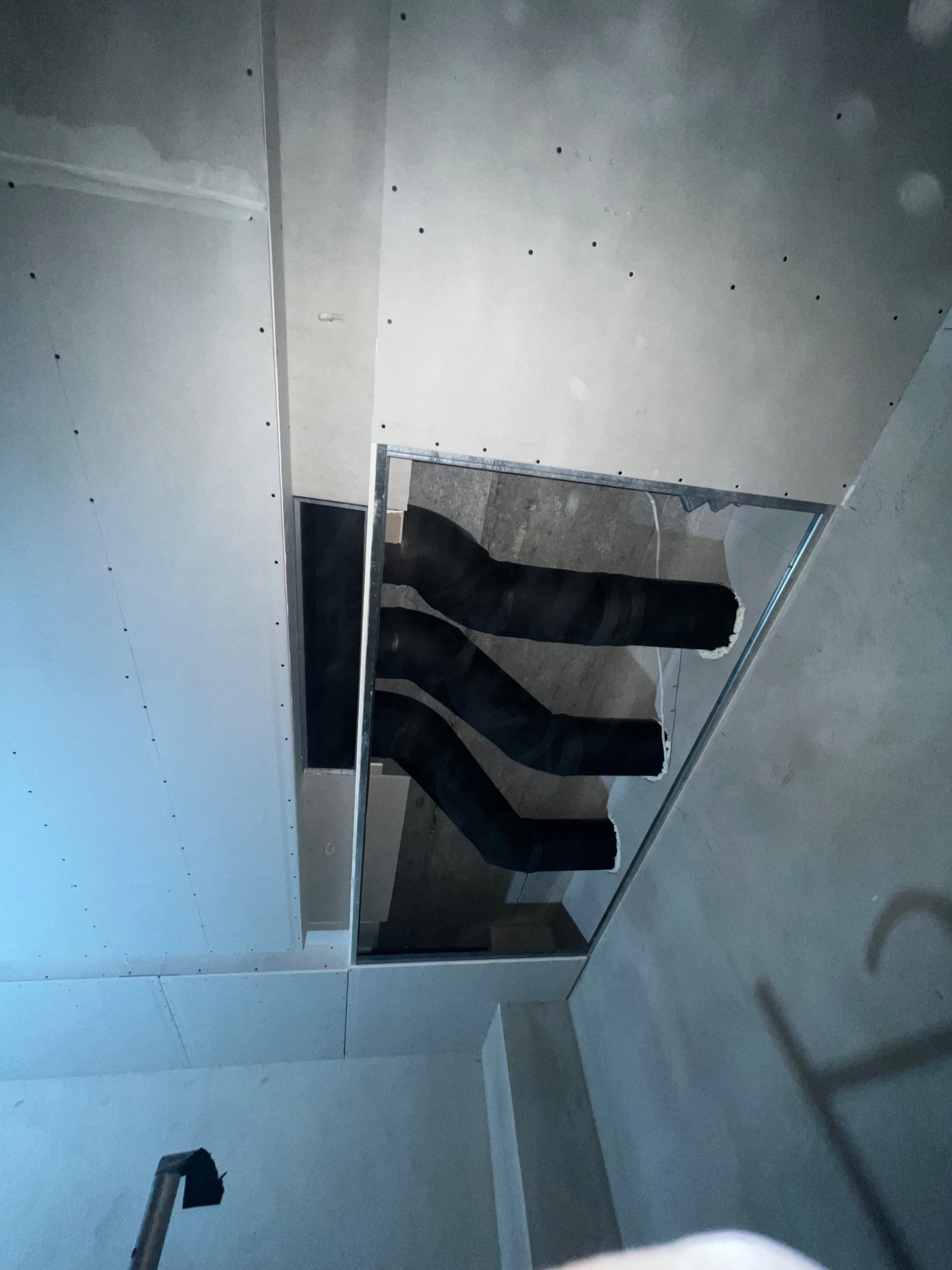 Cztery czarne, zaizolowane rury wentylacyjne widoczne w otworze w suficie podwieszanym z płyt gipsowo-kartonowych, instalacja w trakcie realizacji w Lesznie.