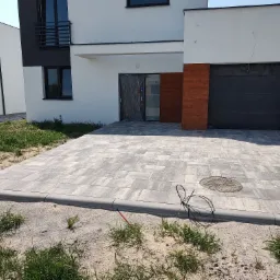 Ułożona szara kostka brukowa na podjeździe nowego domu z szarą bramą garażową i drewnopodobnymi panelami elewacyjnymi; widoczny właz kanalizacyjny i wystające kable elektryczne na trawniku.