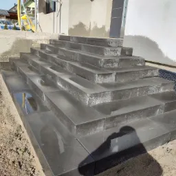 Nowe, ciemnoszare schody z kamiennych bloków prowadzące do domu, widoczne ślady wilgoci na powierzchni stopni, w tle ocieplenie budynku i rusztowanie.