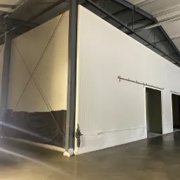 Wnętrze hali z zamontowanymi ścianami z białych płyt warstwowych i dwoma przesuwnymi drzwiami, widoczna stalowa konstrukcja nośna i zabezpieczenia dolnej części ścian czarną folią.