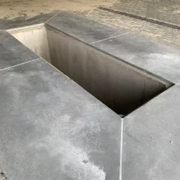 Szary betonowy fundament z otworem inspekcyjnym, widoczne ślady po deskowaniu i faktura betonu, obok kostka brukowa.