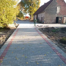 Nowo ułożony chodnik z szarej kostki brukowej z czerwonym obramowaniem prowadzący w kierunku starego, ceglanego budynku i osoby w oddali, z opadłymi liśćmi na powierzchni.