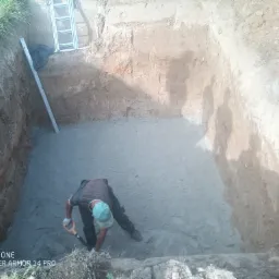 Wykonawca w wykopie wyrównuje podłoże pod budowę, widoczna drabina oparta o ścianę wykopu i poziomica.