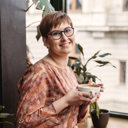 BAFO pracownia - Uśmiechnięta kobieta w okularach, z kolczykiem w wardze, trzymająca filiżankę kawy latte w kawiarni z roślinami w tle.