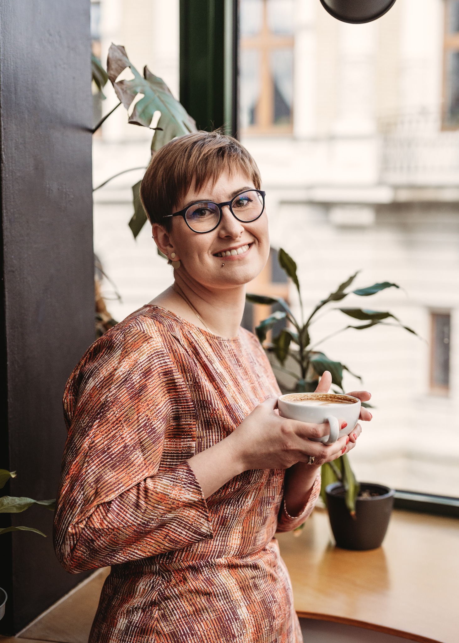 Uśmiechnięta kobieta w okularach, z kolczykiem w wardze, trzymająca filiżankę kawy latte w kawiarni z roślinami w tle.