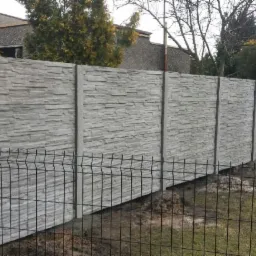 Szary, prefabrykowany mur z betonu imitującego kamień, oddzielający posesję od ulicy, z widoczną siatką ogrodzeniową z przodu.