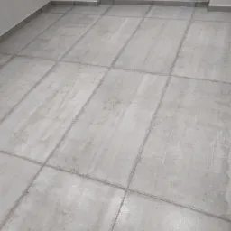 Ułożona jasnoszara podłoga z płytek ceramicznych, widoczne fugi i listwy przypodłogowe, surowy wygląd.