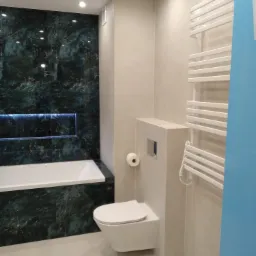 Nowoczesna łazienka z wanną obudowaną płytkami imitującymi ciemny marmur z podświetlaną wnęką, podwieszaną toaletą i grzejnikiem drabinkowym.