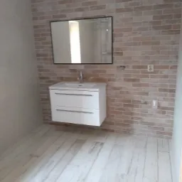 Wykończona łazienka z umywalką na szafce, lustrem w czarnej ramie i ścianami wyłożonymi płytkami imitującymi cegłę w odcieniach beżu i brązu, kremowa podłoga z płytek imitujących drewno.