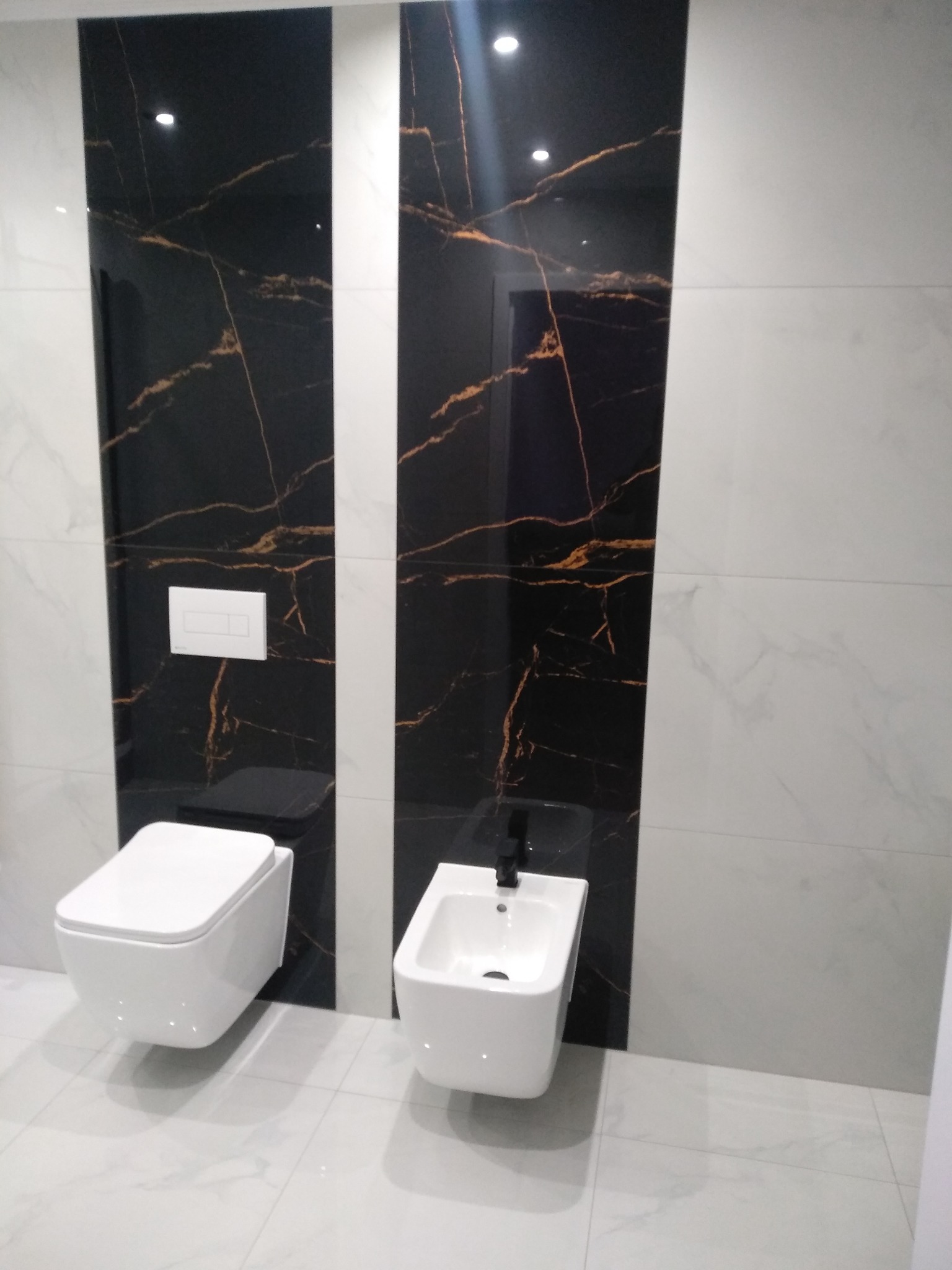 Nowoczesna łazienka z białą wiszącą toaletą i bidetem na tle czarno-białych płytek imitujących marmur ze złotymi żyłami, widoczny przycisk spłuczki na ścianie.