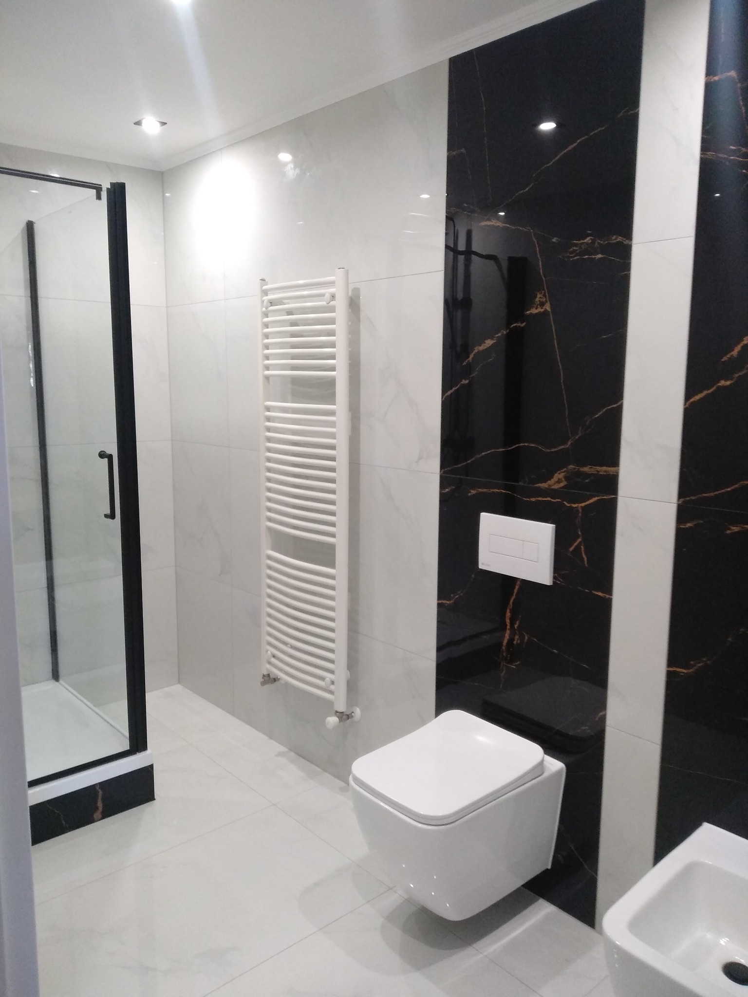 Nowoczesna łazienka z kabiną prysznicową z czarną ramą, grzejnikiem drabinkowym, wiszącą toaletą i umywalką, wykończona białymi i czarnymi płytkami imitującymi marmur.