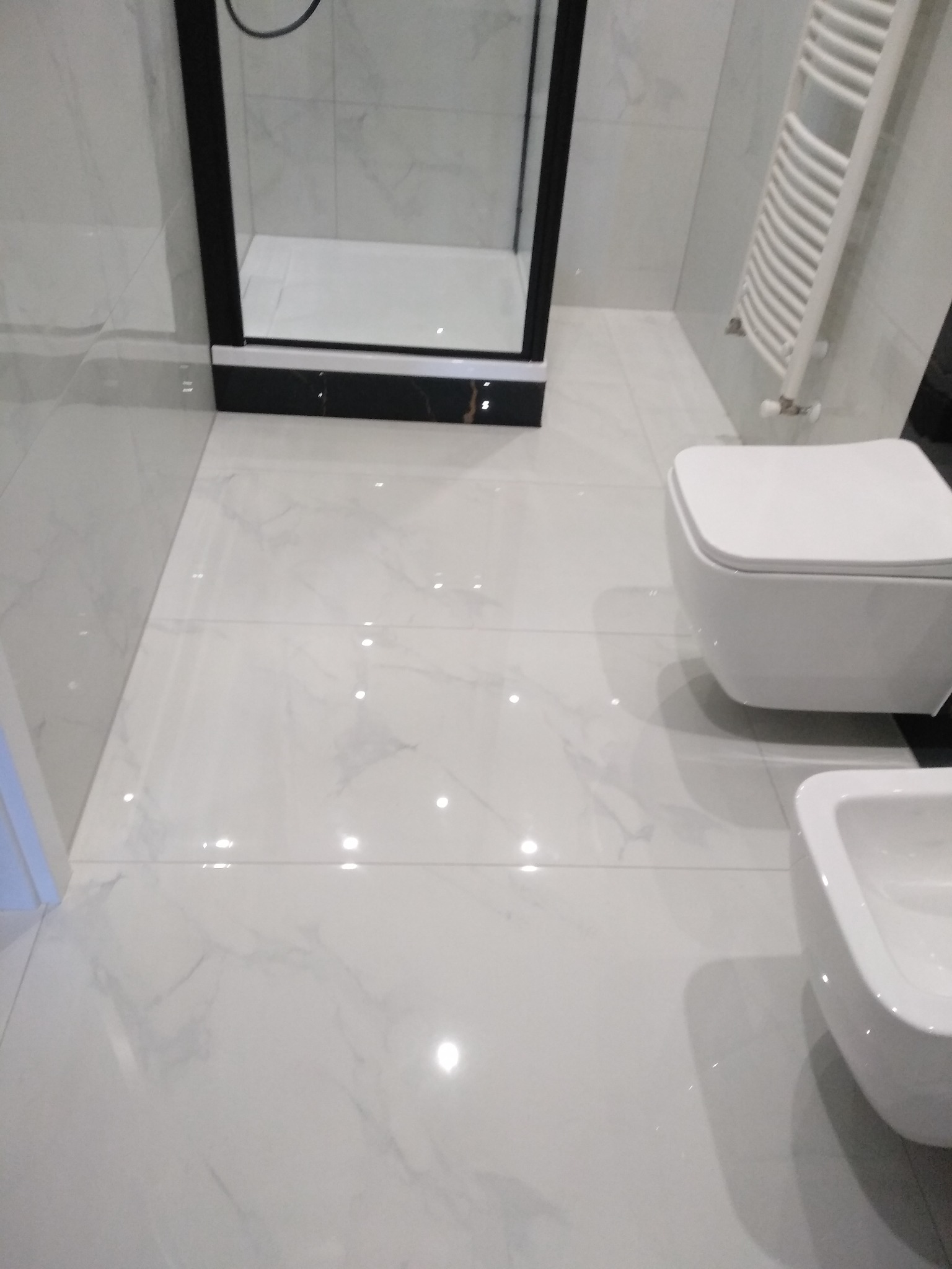 Biała łazienka z kabiną prysznicową z czarną ramą, podwieszaną toaletą i bidetem, wykończona płytkami imitującymi marmur.