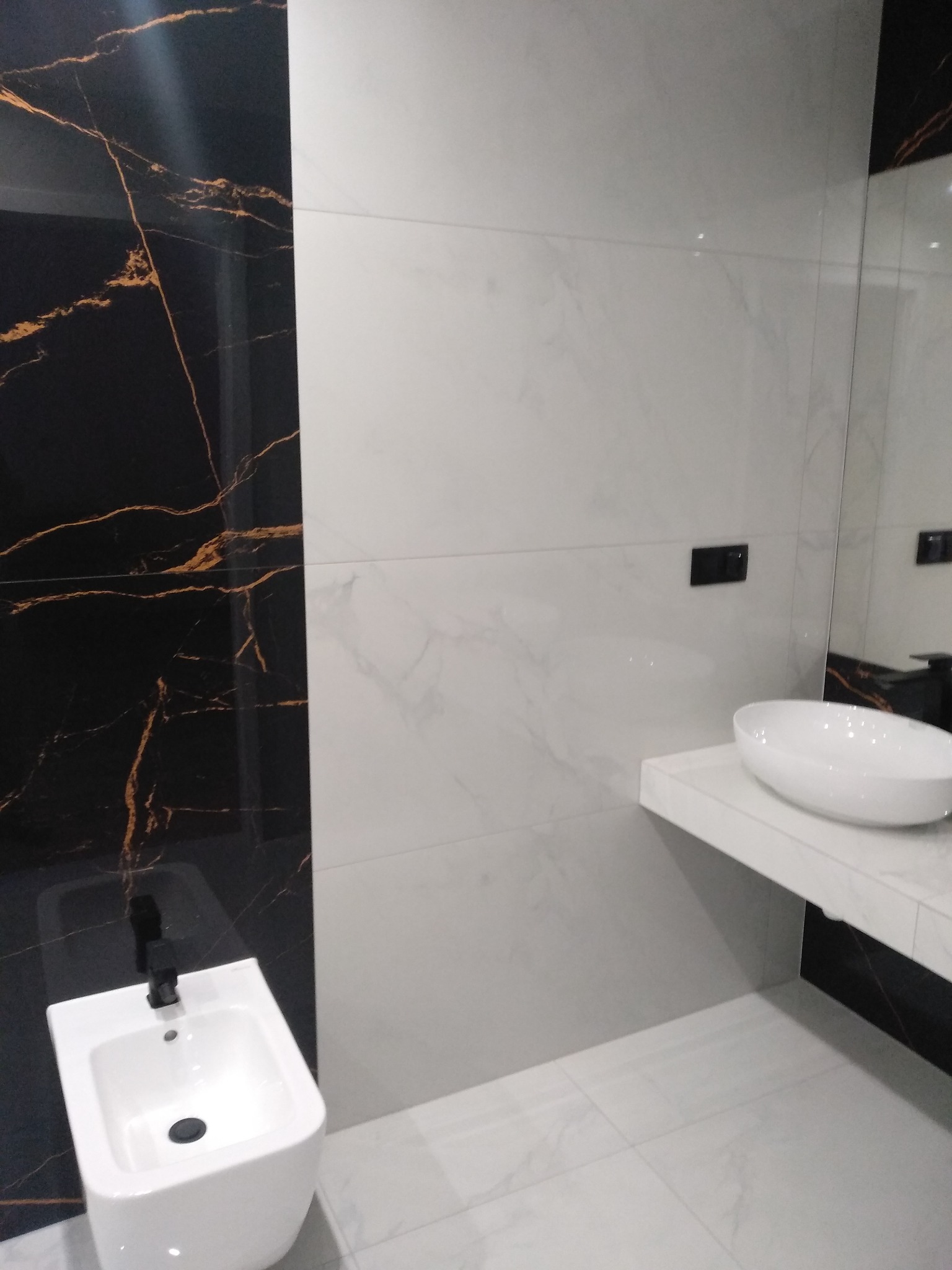 Nowoczesna łazienka z czarno-złotą płytką marmurową, białą umywalką nablatową i bidetem z czarną armaturą.
