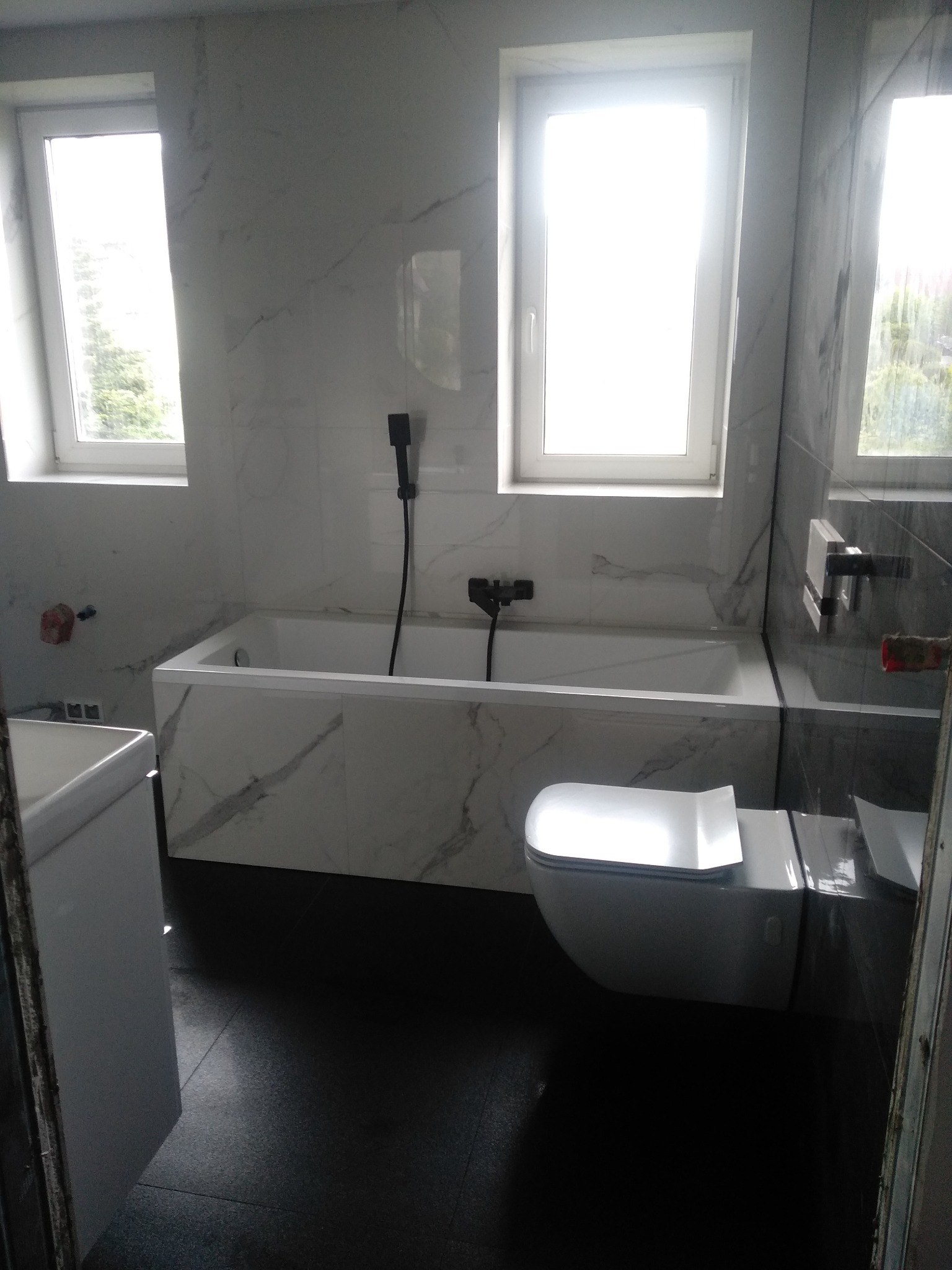 Nowoczesna łazienka z wanną obudowaną płytami imitującymi marmur, wiszącą miską WC i czarną armaturą, wykończona płytkami w odcieniach szarości i bieli.