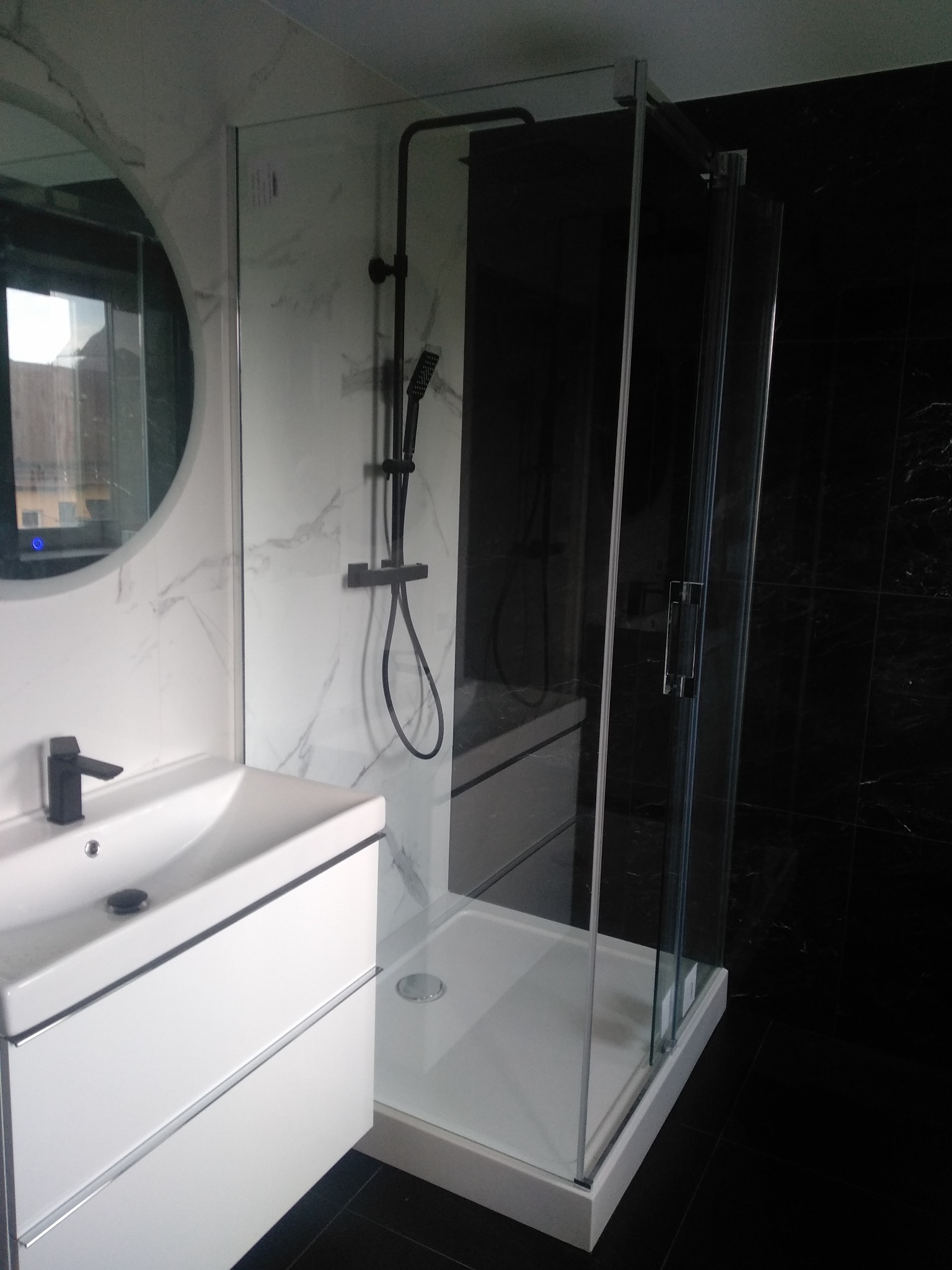 Nowoczesna łazienka z kabiną prysznicową typu walk-in, umywalką z szafką w kolorze białym i czarną armaturą na tle białych i czarnych płytek imitujących marmur.