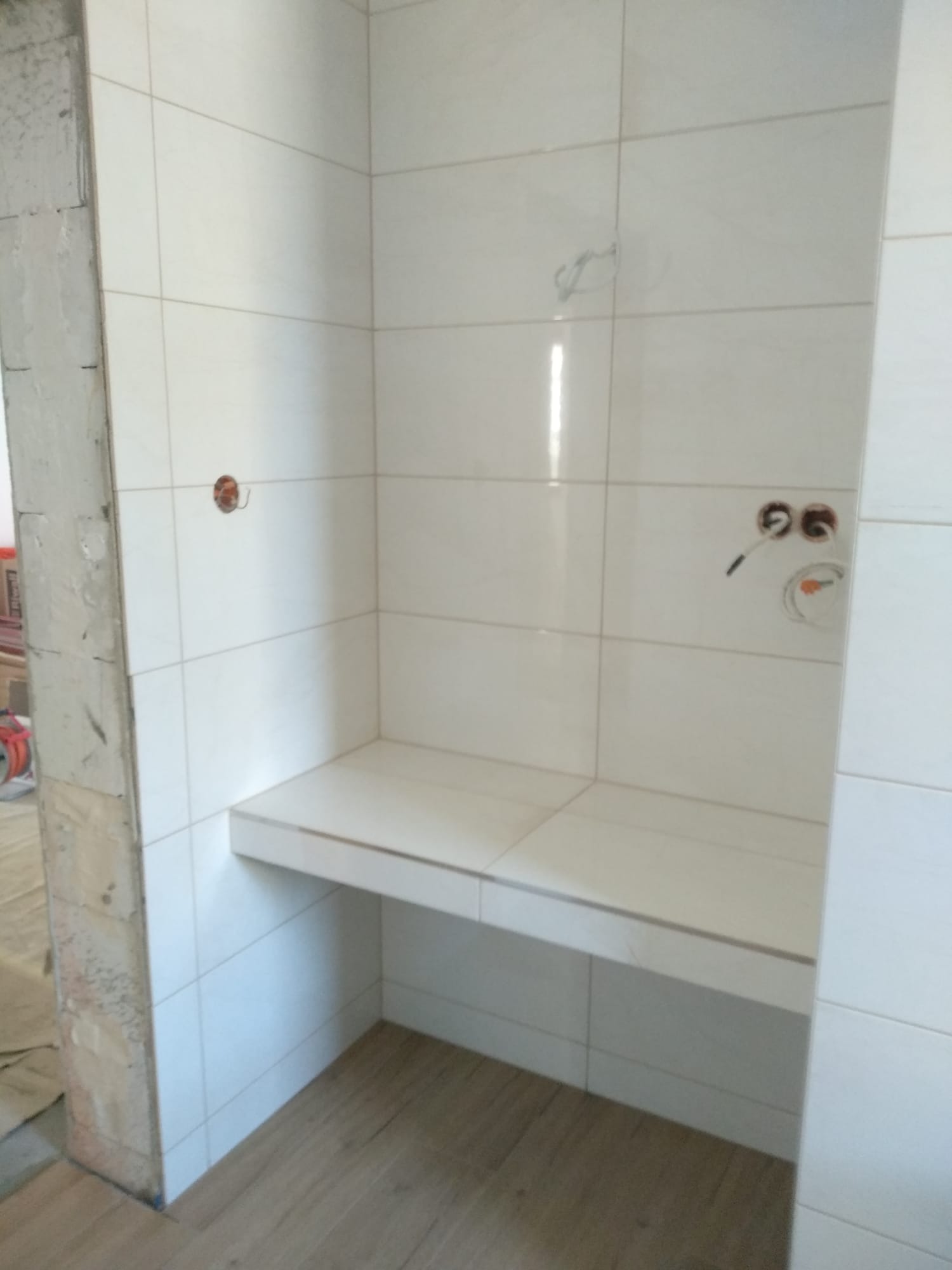 Wnęka prysznicowa w trakcie wykańczania, z obłożonymi białymi płytkami ścianami i półką, widoczne przygotowane otwory na armaturę, surowy fragment ściany obok.