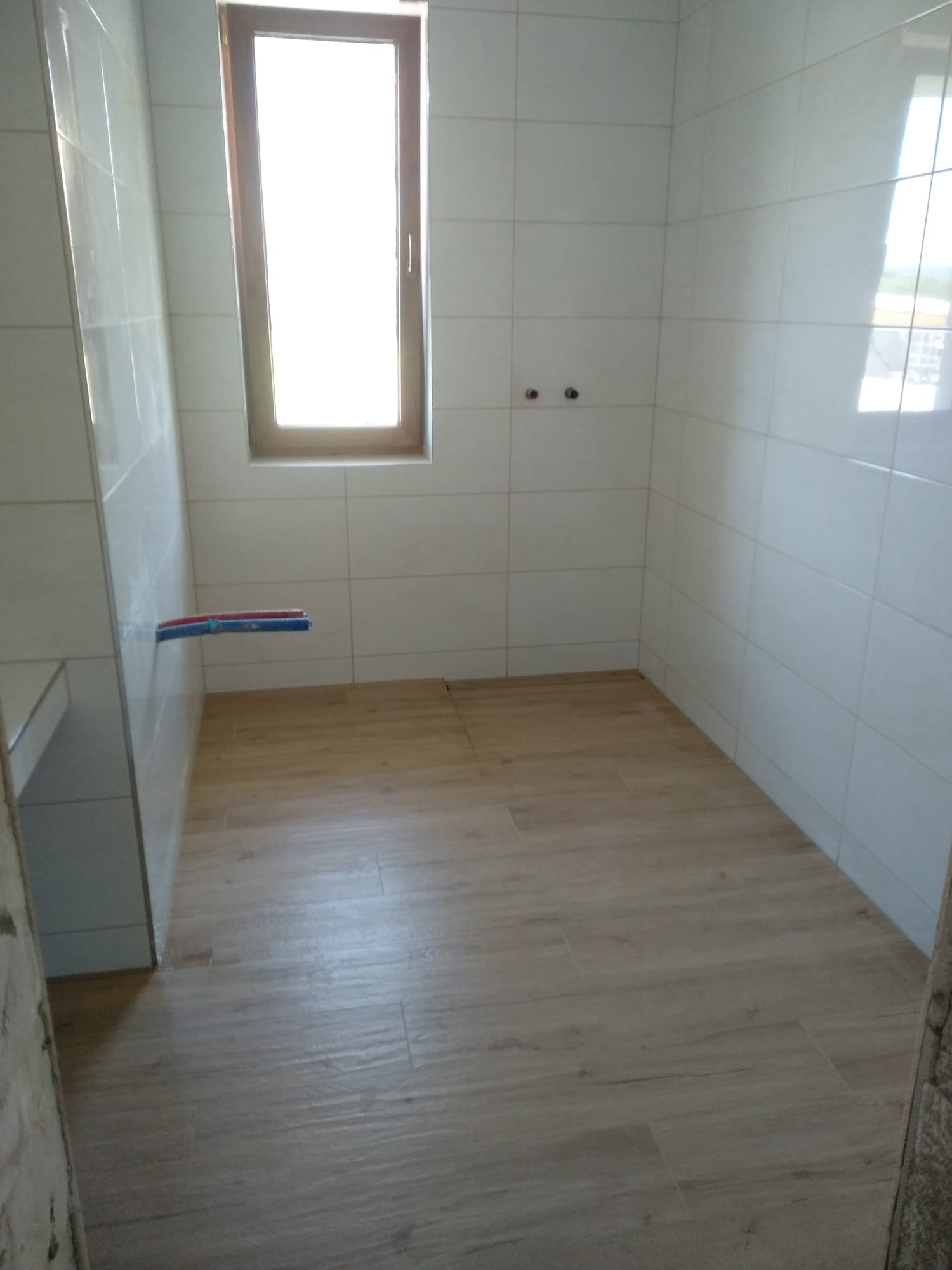 Wykończona łazienka z beżowymi płytkami podłogowymi imitującymi drewno, białymi płytkami ściennymi, oknem w drewnianej ramie i wystającymi rurami instalacji wodnej.