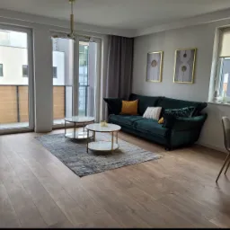 Jasne wnętrze salonu z parkietem, ciemnozieloną sofą z poduszkami, okrągłymi stolikami kawowymi na dywanie i oknami balkonowymi z szarymi zasłonami.