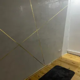 Szara ściana z dekoracyjnymi, złotymi paskami tworzącymi geometryczny wzór, drewniana podłoga i czarny dywanik.