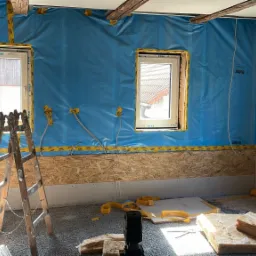 Wnętrze remontowanego poddasza z widoczną izolacją ścian folią, oknami z żółtą taśmą zabezpieczającą, fragmentami płyt OSB i rozłożoną wełną mineralną na podłodze, z drabinką opartą o ścianę.