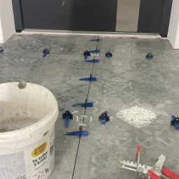 Układanie szarych płytek podłogowych przy użyciu systemu poziomowania, widoczny biały pojemnik z klejem, narzędzie do dociskania płytek z czerwonym uchwytem, sznurek traserski i fragment czerwonej...