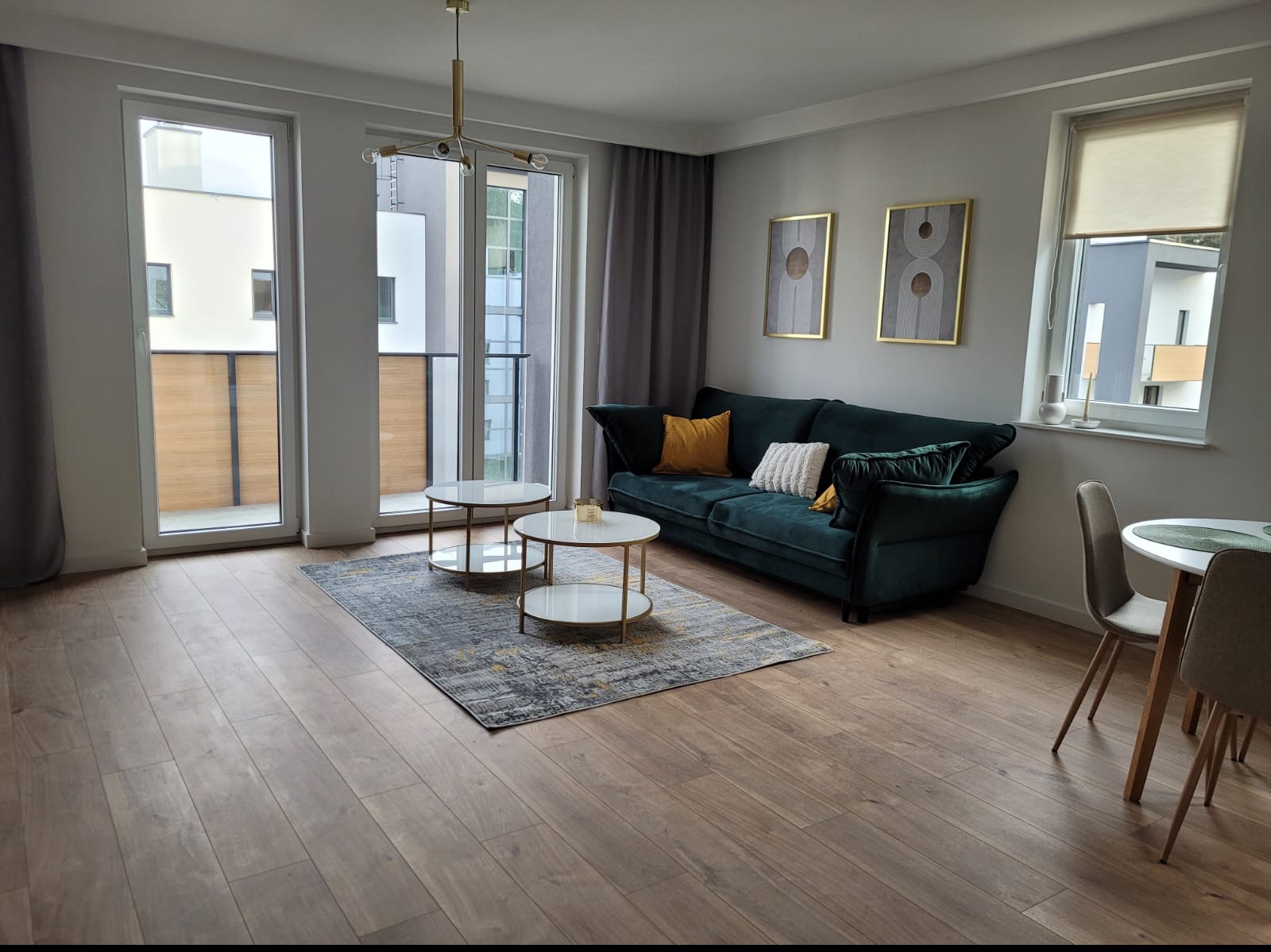 Jasne wnętrze salonu z parkietem, ciemnozieloną sofą z poduszkami, okrągłymi stolikami kawowymi na dywanie i oknami balkonowymi z szarymi zasłonami.