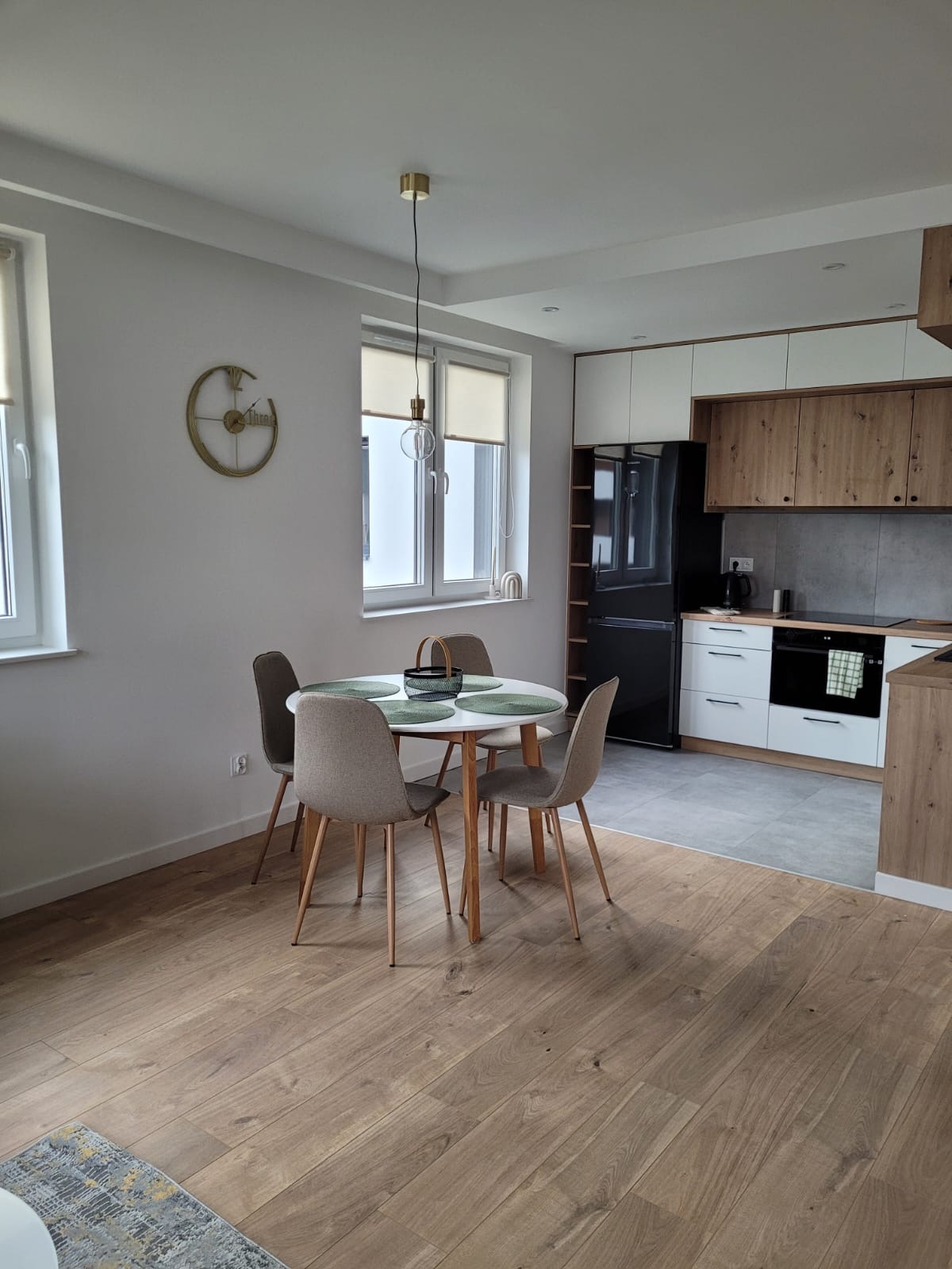 Jasne wnętrze mieszkania z drewnianą podłogą, okrągłym stołem jadalnym z czterema krzesłami i nowoczesną zabudową kuchenną z białymi i drewnianymi frontami, czarną lodówką i szarą ścianą nad blatem.