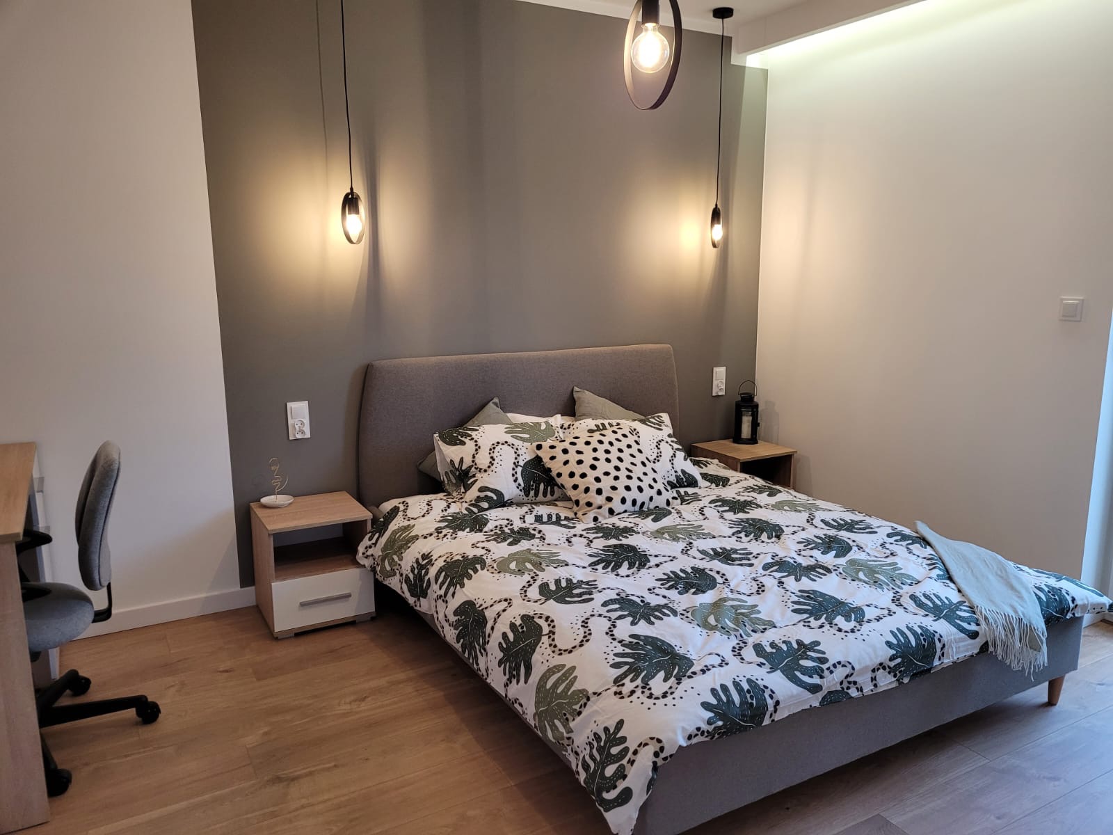 Nowoczesna sypialnia z łóżkiem z szarą ramą i pościelą w liście, oświetlona wiszącymi lampami z ciepłym światłem, na drewnianej podłodze.
