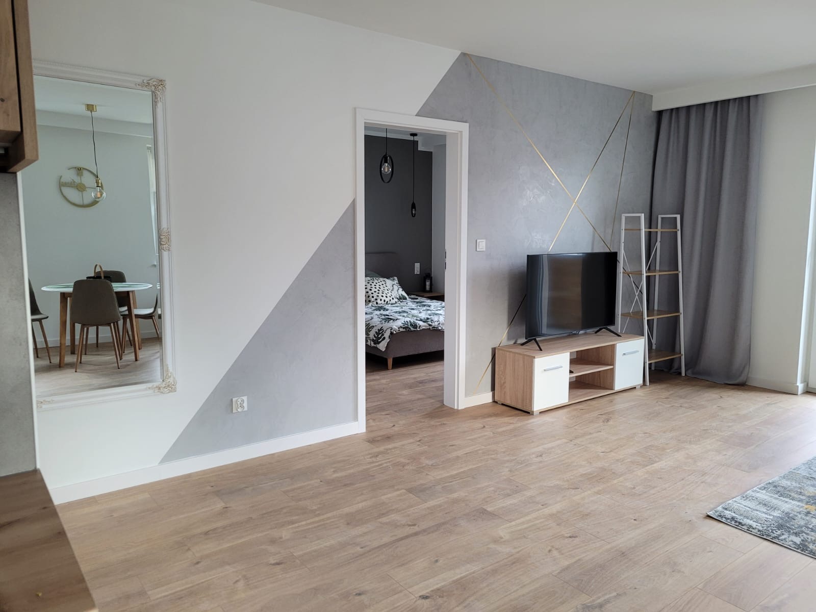 Pokój dzienny po remoncie: ściana z dekoracyjną szpachlą w odcieniach szarości i bieli, zdobiona złotymi liniami, drewniana podłoga, telewizor na szafce RTV, w tle widok na sypialnię przez otwarte...