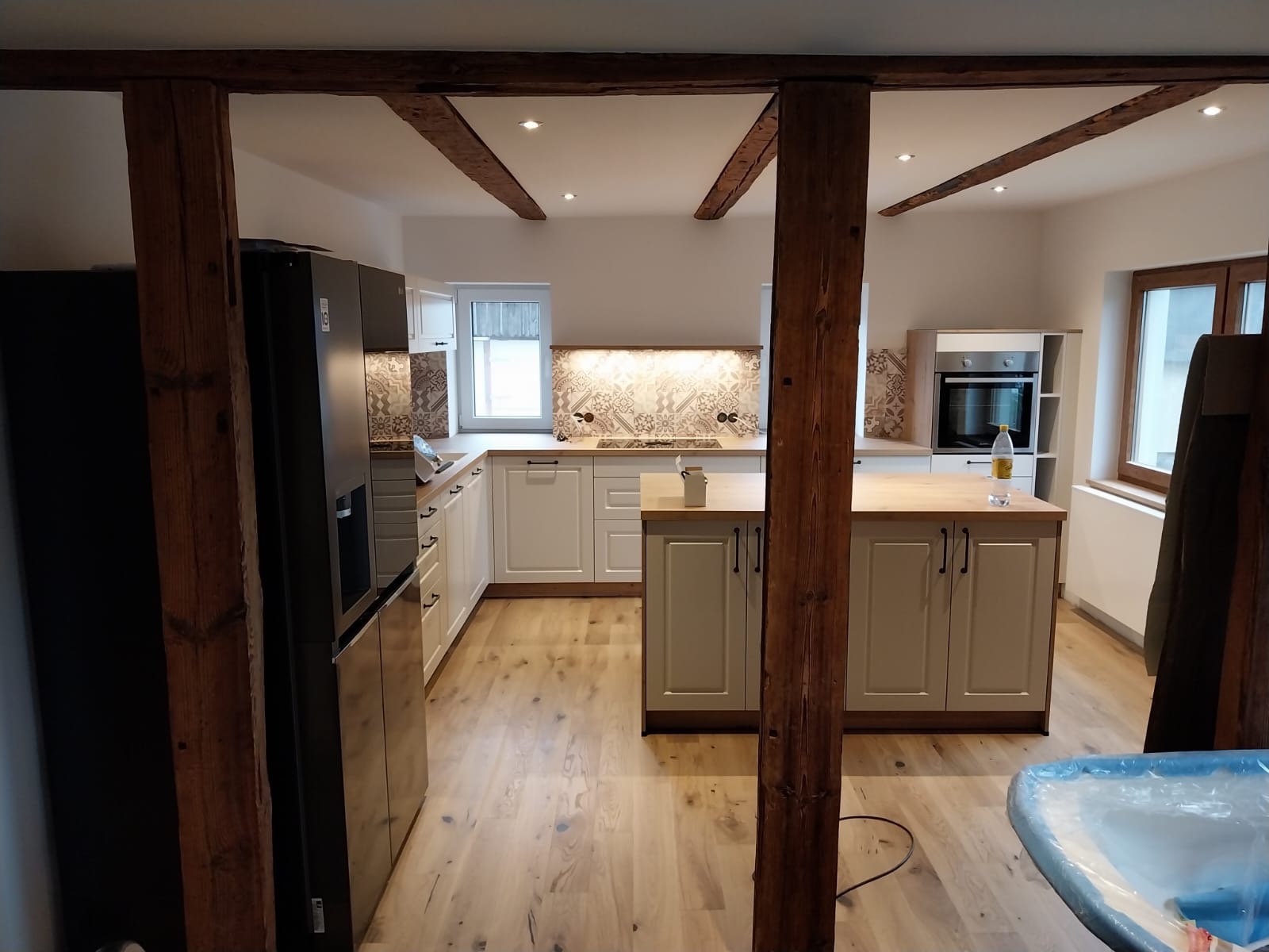 Wykończona kuchnia w stylu rustykalnym z drewnianymi belkami stropowymi, jasnymi szafkami i wyspą, wzorzystą glazurą nad blatem i czarną lodówką z dozownikiem wody.