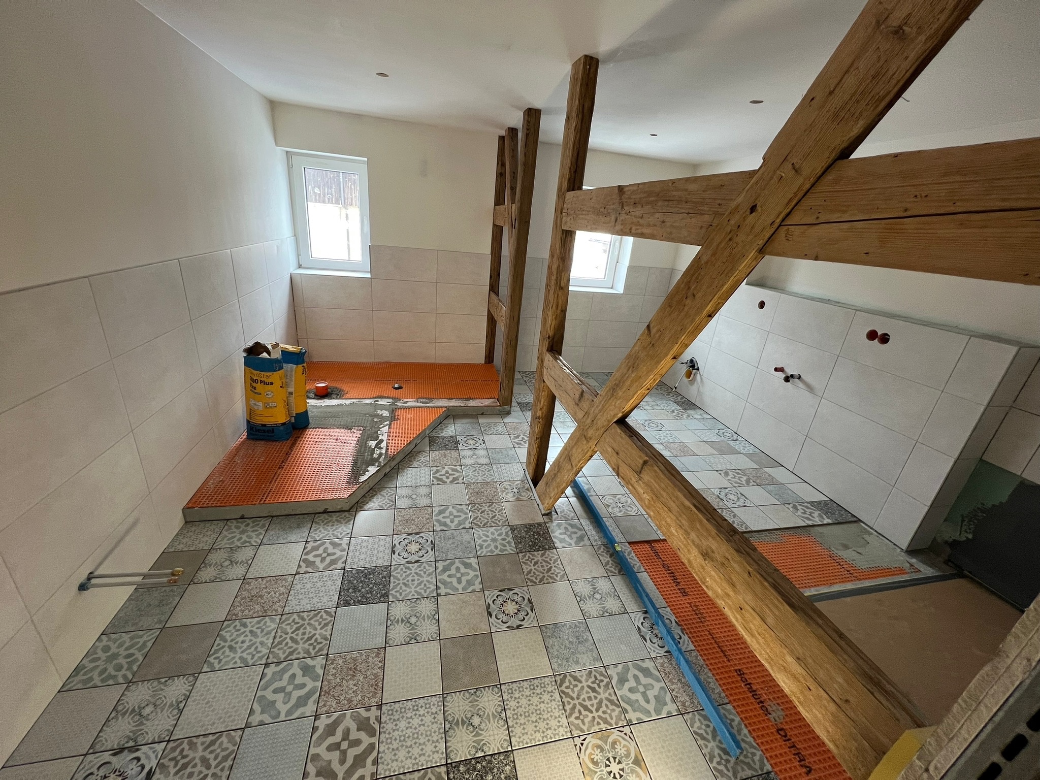 Remontowana łazienka z drewnianą konstrukcją stropu, podłoga w patchworkowe płytki, fragment podłogi z pomarańczową matą izolacyjną, dwa worki kleju do płytek przy ścianie.