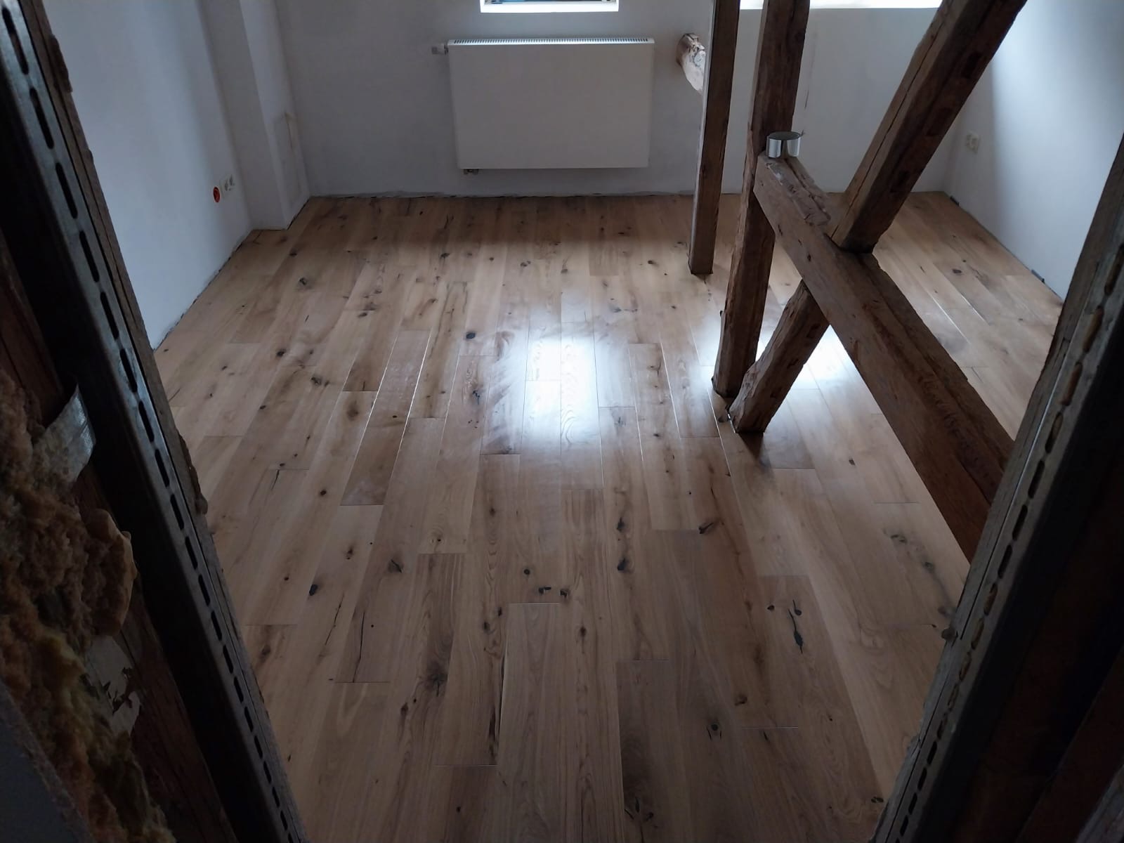 Jasna, drewniana podłoga z wyraźnym rysunkiem słojów, ułożona w odnowionym poddaszu z widocznymi elementami drewnianej konstrukcji dachu i fragmentem ocieplenia.