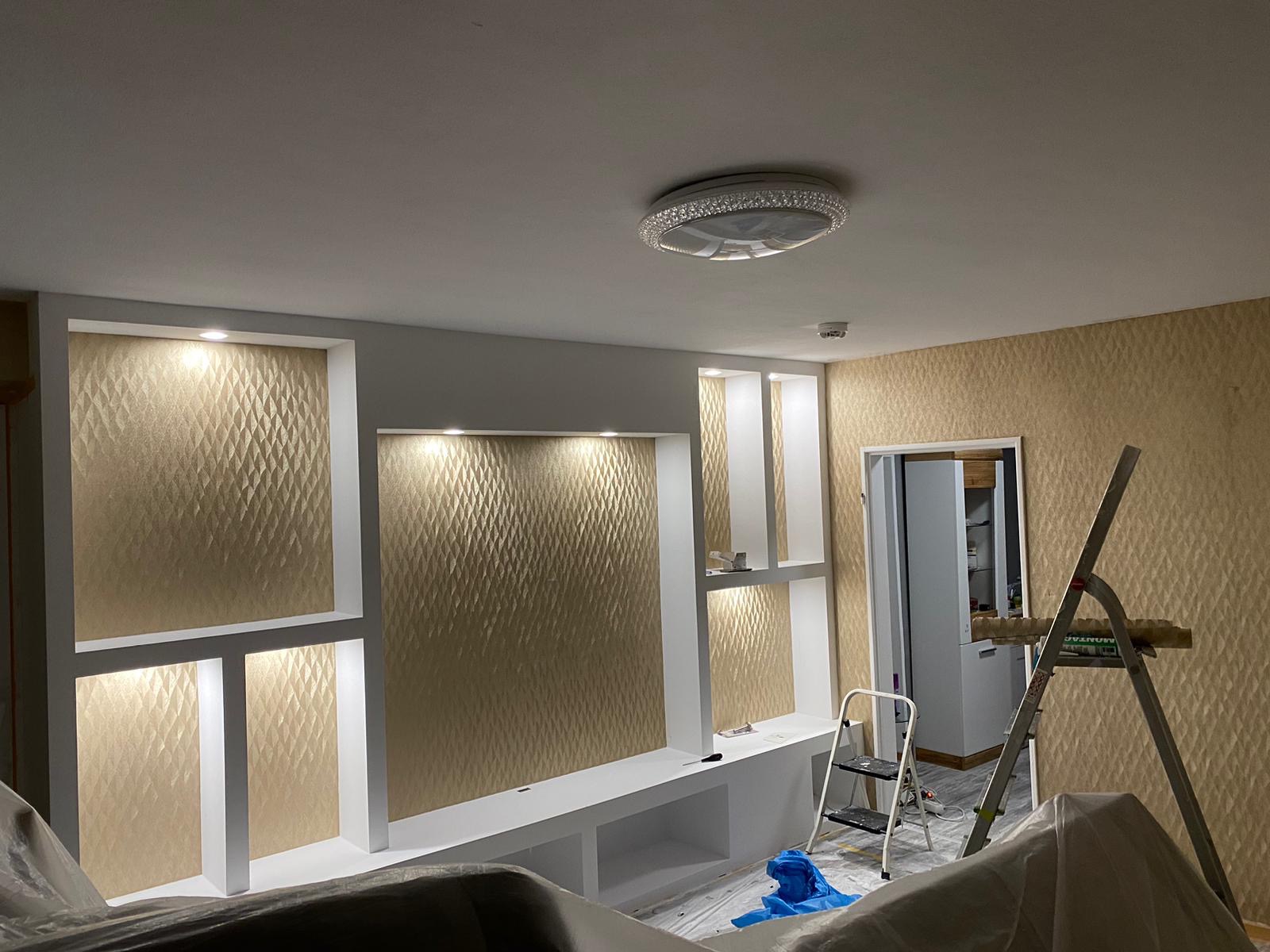 Wykończone wnętrze z dekoracyjną ścianą z wnękami oświetlonymi punktowo, widoczna drabina malarska i folia ochronna na meblach.