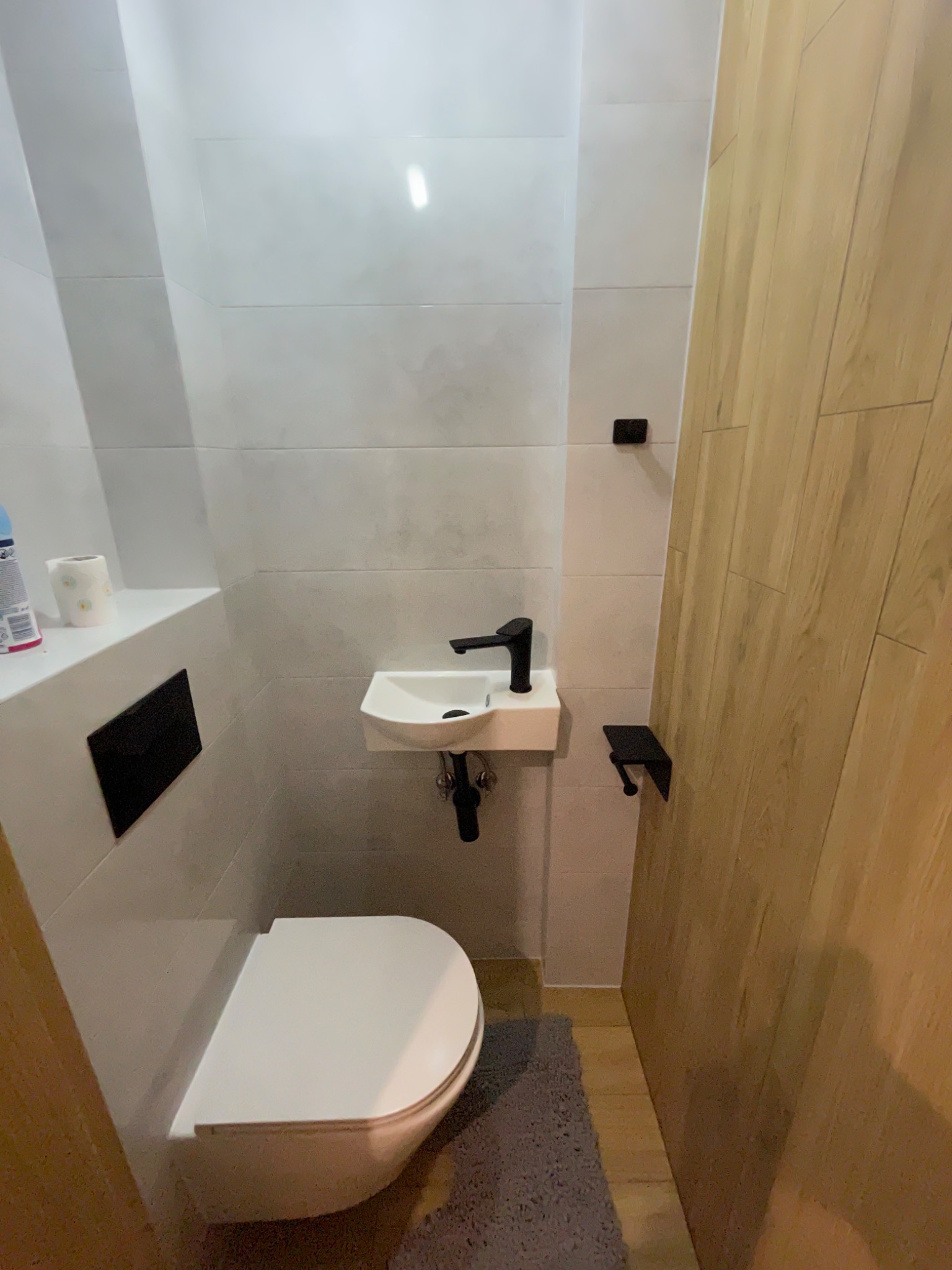 Mała łazienka z wiszącą toaletą i umywalką, czarna armatura, szare płytki ścienne i drewniane panele na drzwiach, widoczny dywanik.