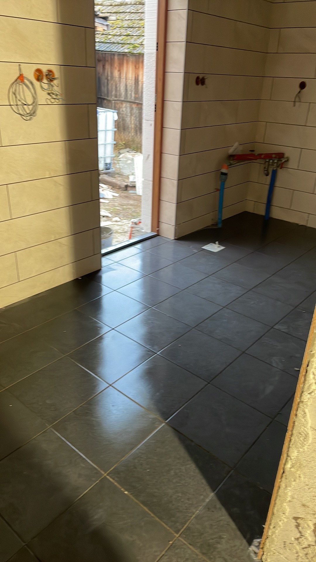 Wnętrze łazienki w trakcie remontu, widoczne świeżo położone ciemne płytki podłogowe, jasne płytki ścienne, wystające kable elektryczne i instalacja hydrauliczna z niebieskimi rurami.