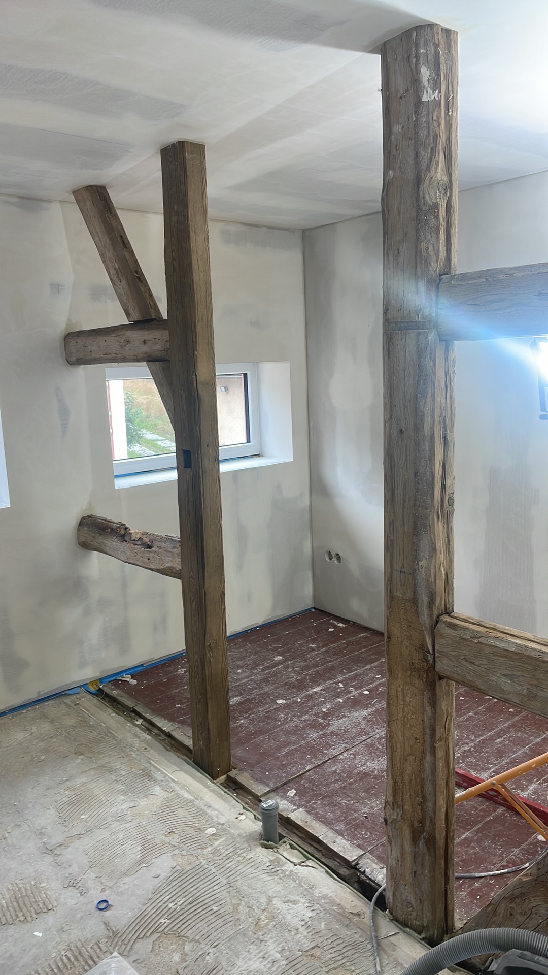 Wnętrze w trakcie remontu: drewniane belki konstrukcyjne, częściowo wyszpachlowane ściany, drewniana podłoga z widocznymi śladami prac, okno z widokiem na zieleń.