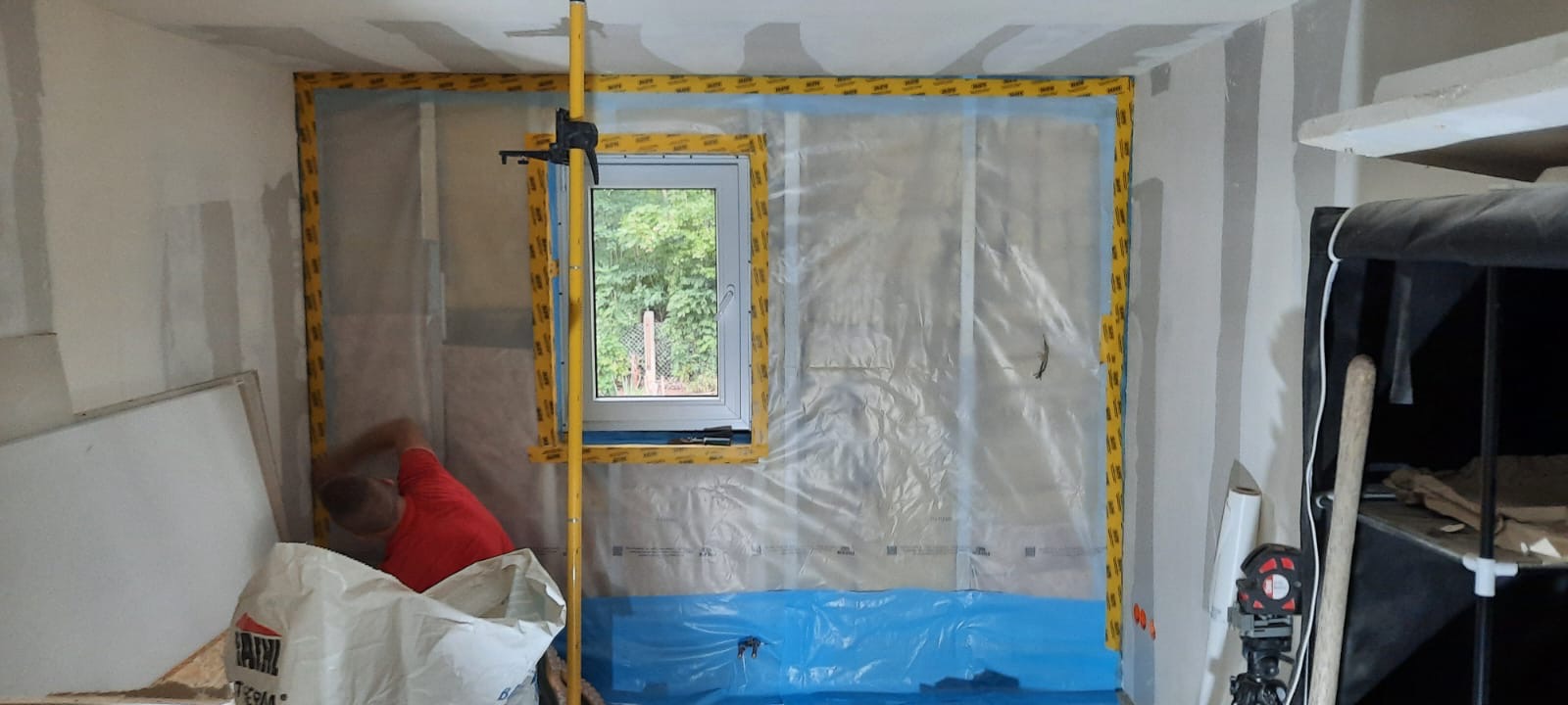 Montaż folii ochronnej na ścianie z oknem w Biłgoraju, przygotowanie do prac wykończeniowych, widoczny pracownik w czerwonej koszulce i narzędzia pomiarowe.