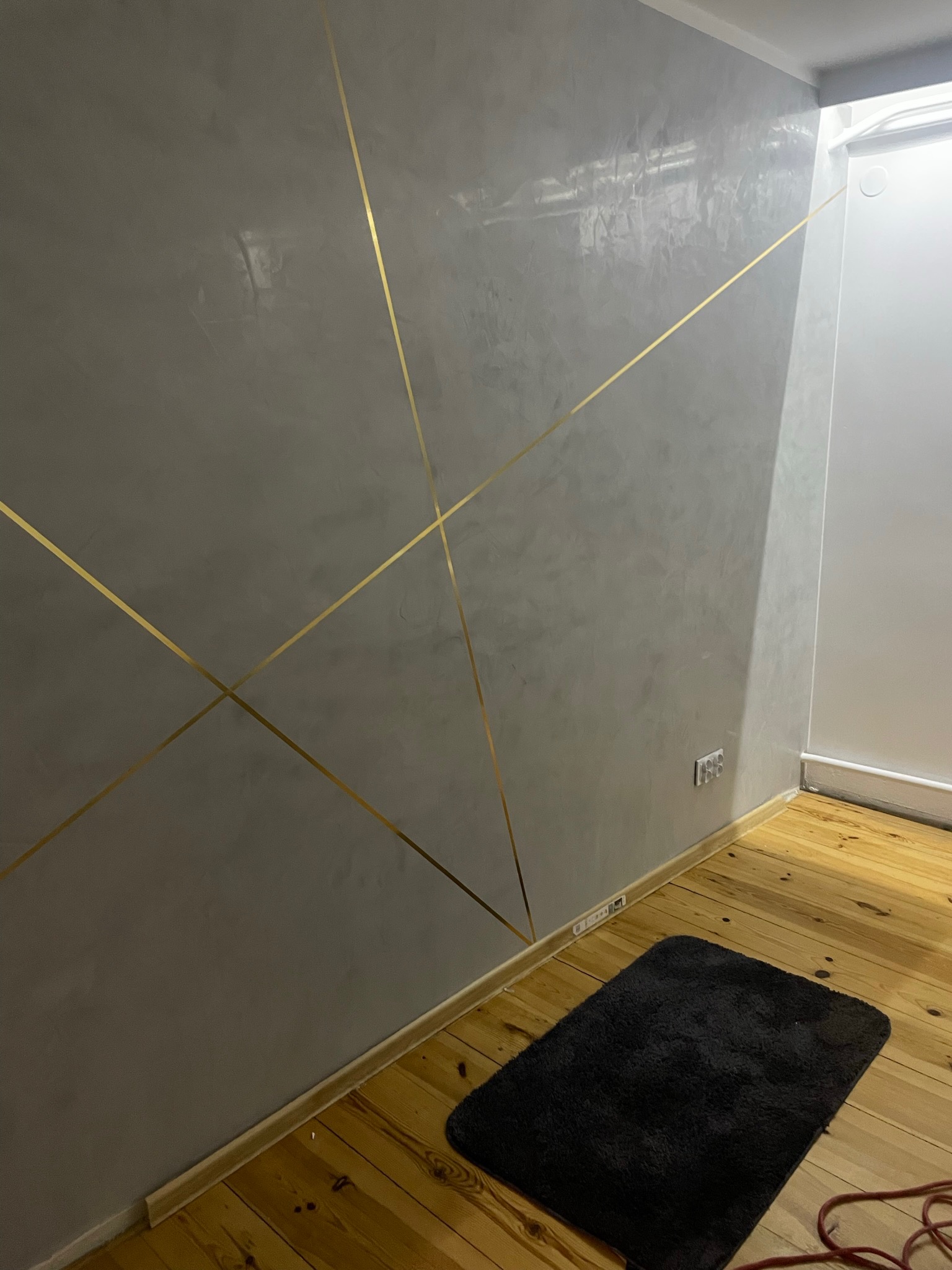 Szara ściana z dekoracyjnymi, złotymi paskami tworzącymi geometryczny wzór, drewniana podłoga i czarny dywanik.
