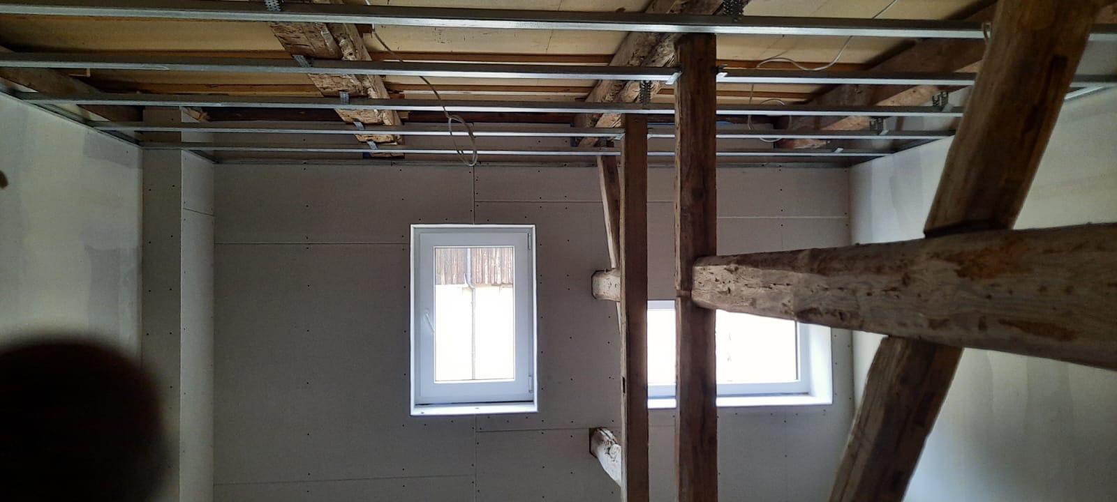 Wnętrze remontowanego poddasza z widocznymi drewnianymi belkami konstrukcyjnymi, zamontowaną metalową konstrukcją sufitu podwieszanego i ścianami wyłożonymi płytami. Dwa okna w białych ramach w tle.