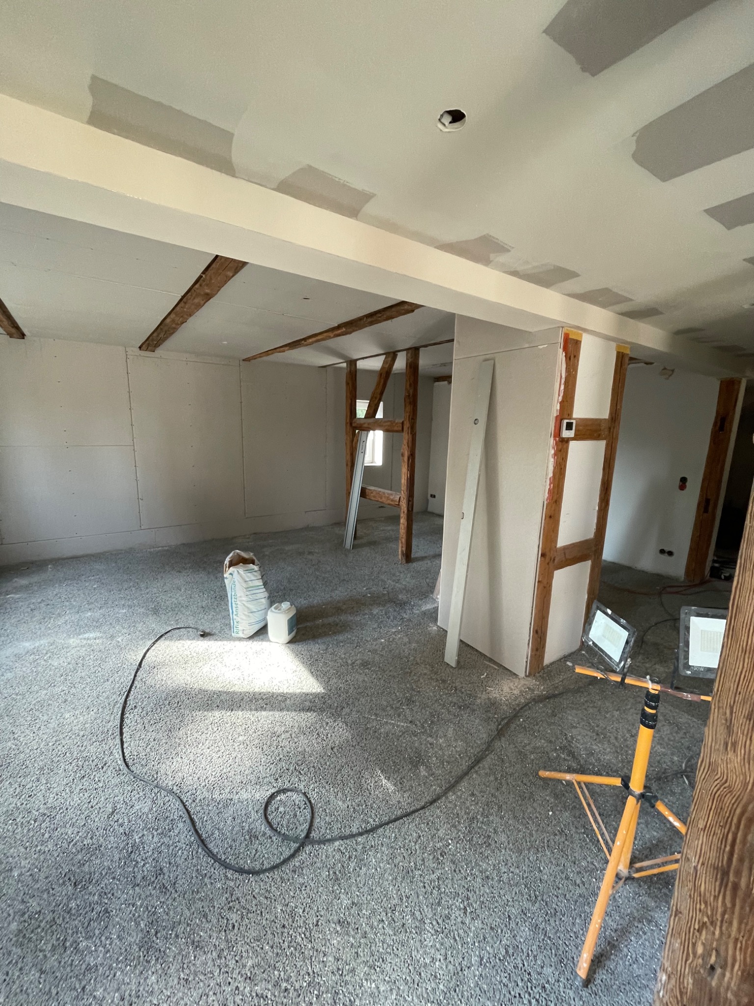 Wnętrze w trakcie prac wykończeniowych: surowy strop z drewnianymi belkami, ściany częściowo pokryte płytami, worki z materiałami budowlanymi na podłodze, oświetlenie budowlane na statywach.