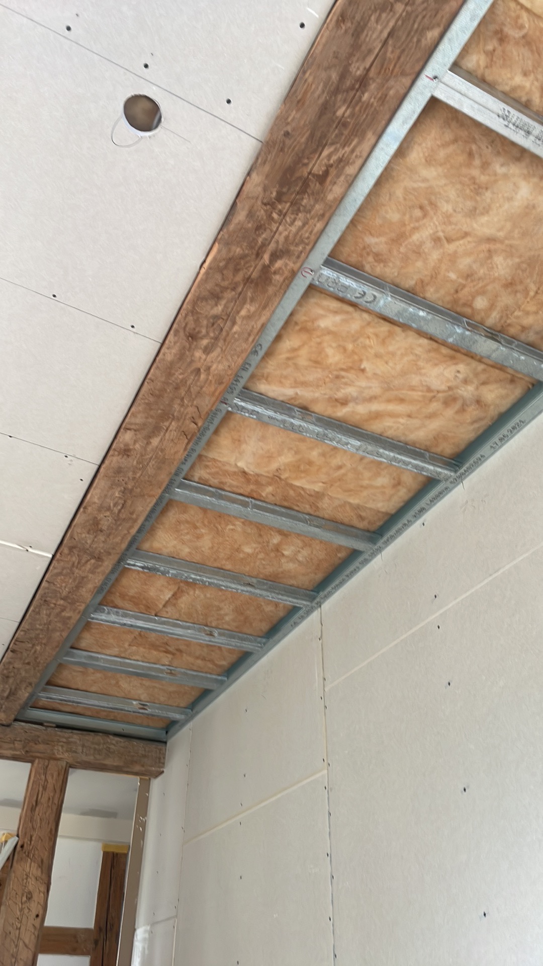 Konstrukcja metalowa podwieszanego sufitu z izolacją z wełny mineralnej, częściowo zakryta płytami gipsowo-kartonowymi, widoczna drewniana belka stropowa i fragment drabiny.