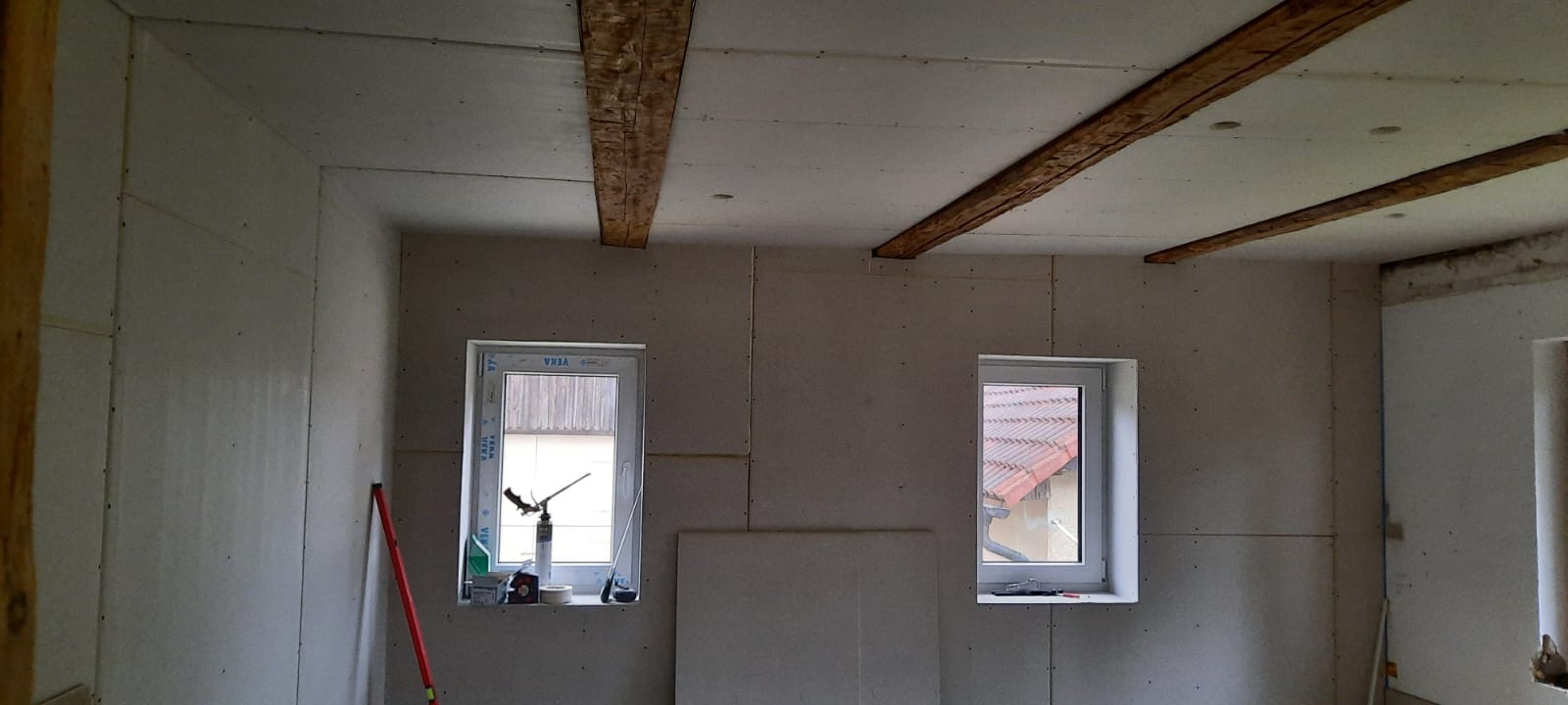 Pokój w trakcie wykańczania płytami karton gips, widoczne drewniane belki stropowe, dwa okna z widokiem na zewnątrz, czerwona poziomica oparta o ścianę.