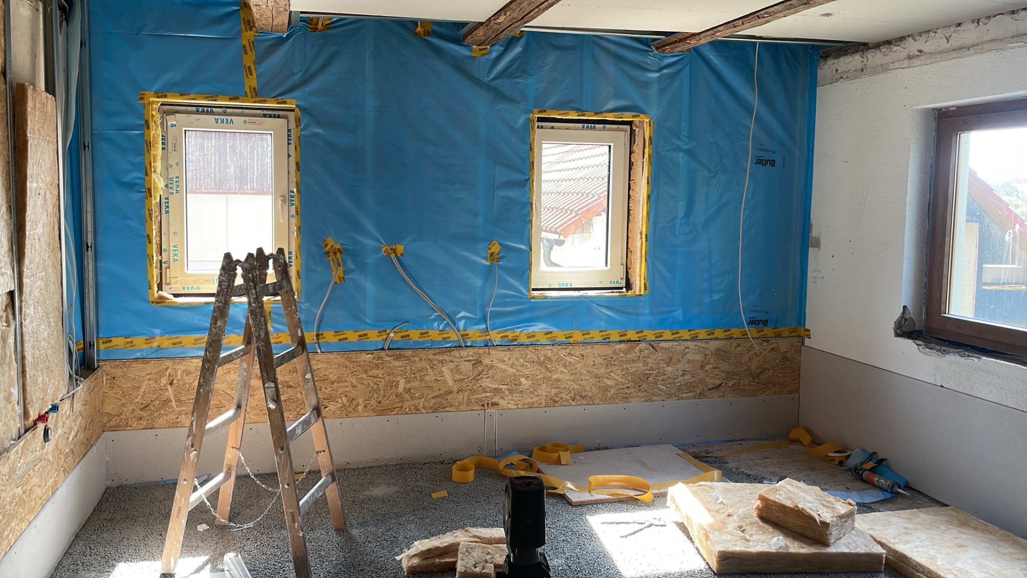 Wnętrze remontowanego poddasza z widoczną izolacją ścian folią, oknami z żółtą taśmą zabezpieczającą, fragmentami płyt OSB i rozłożoną wełną mineralną na podłodze, z drabinką opartą o ścianę.
