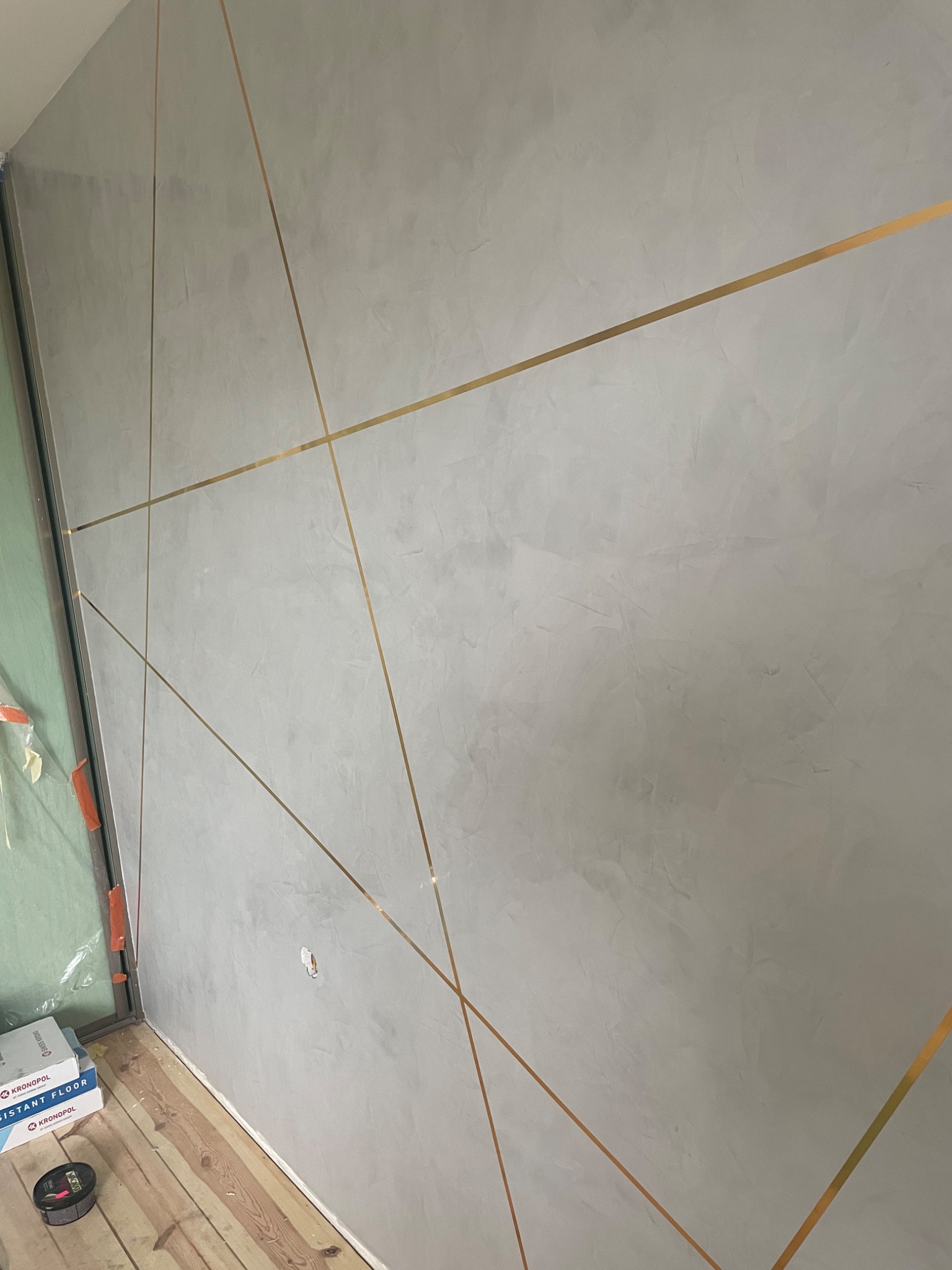 Ściana pokryta szarą dekoracyjną fakturą z geometrycznym wzorem z cienkich, złotych pasków, widoczny fragment podłogi z drewnianych desek i opakowania paneli podłogowych.