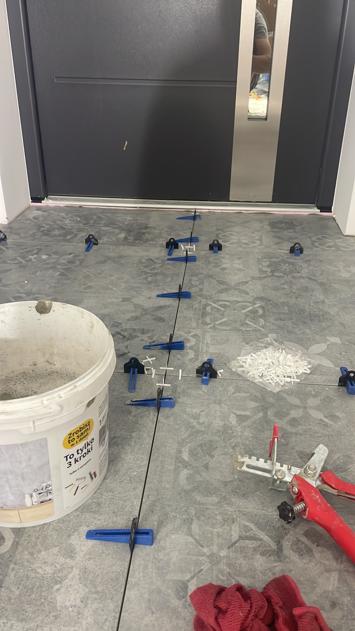 Układanie szarych płytek podłogowych przy użyciu systemu poziomowania, widoczny biały pojemnik z klejem, narzędzie do dociskania płytek z czerwonym uchwytem, sznurek traserski i fragment czerwonej...