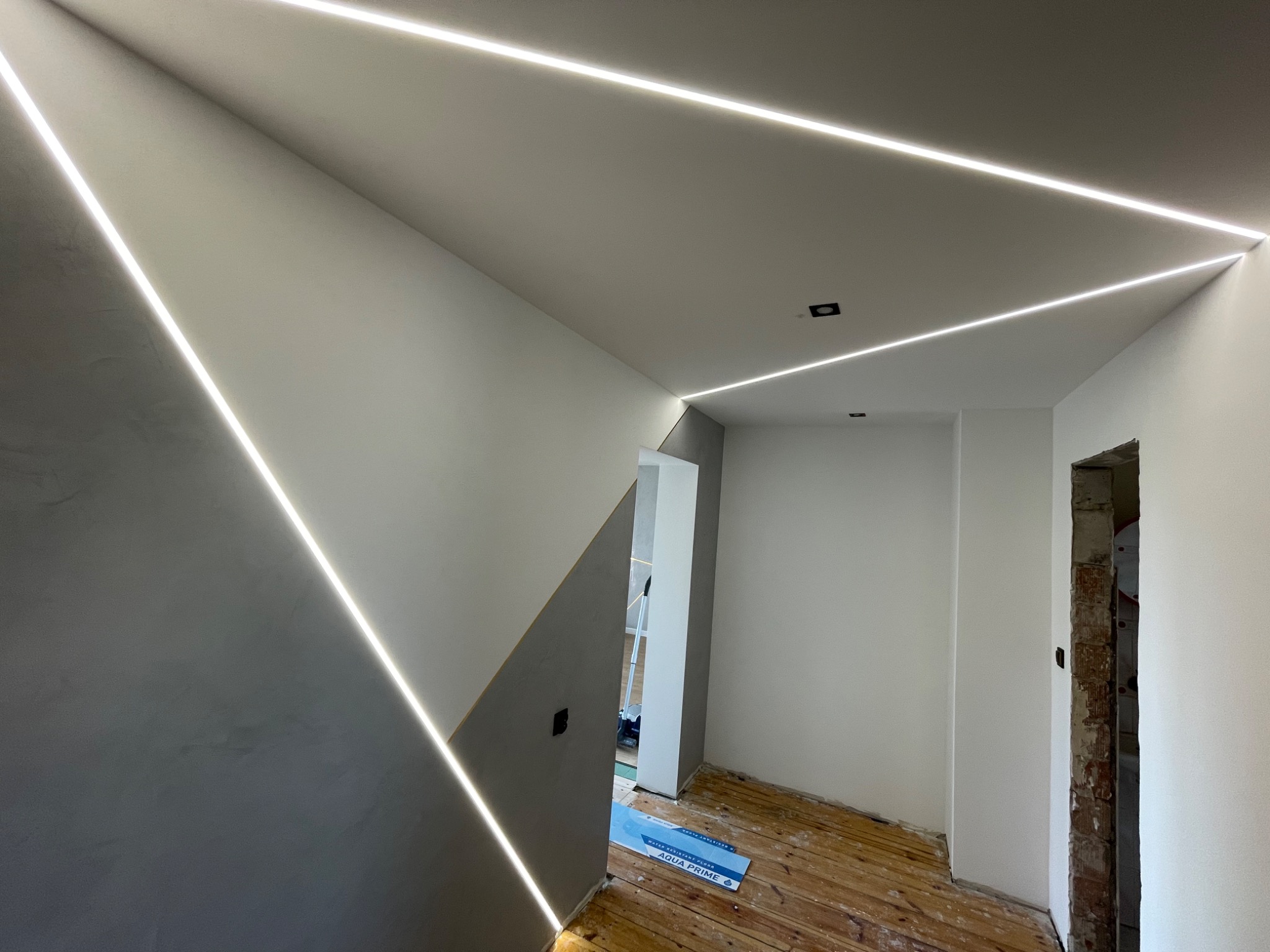 Nowoczesne oświetlenie LED w kształcie trójkąta na suficie i ścianach, szara ściana z efektem dekoracyjnym, drewniana podłoga w trakcie remontu, widoczne ślady prac wykończeniowych.