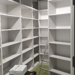 Białe, puste szafy wnękowe z p&oacute;łkami, widoczna drabina i narzędzia na zielonej wykładzinie, sugerujące etap montażu.