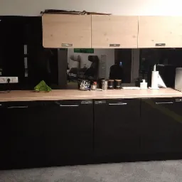 Nowoczesna zabudowa kuchenna z czarnymi, błyszczącymi frontami szafek dolnych i jasnymi szafkami górnymi, uzupełniona o ekspres do kawy, czajnik elektryczny i inne akcesoria kuchenne na blacie.