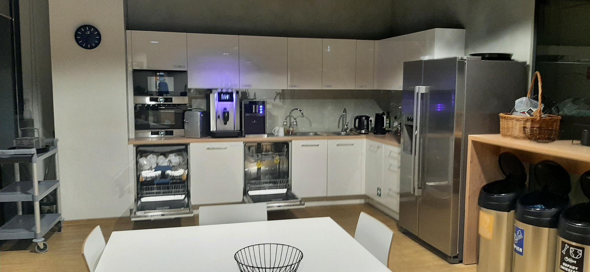 Nowoczesna, biała zabudowa kuchenna z dwoma otwartymi zmywarkami, ekspresami do kawy, lodówką ze stali nierdzewnej i koszami do segregacji śmieci. Na ścianie wisi okrągły zegar.